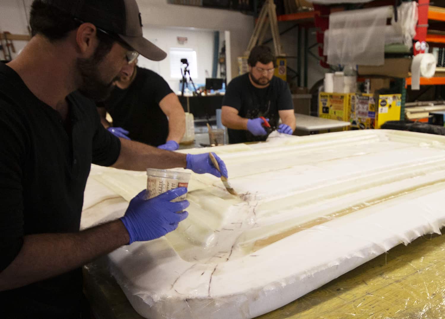 3 men in black t-shirts and purple gloves applying epoxy to fiberglass sheets on a foam mold.